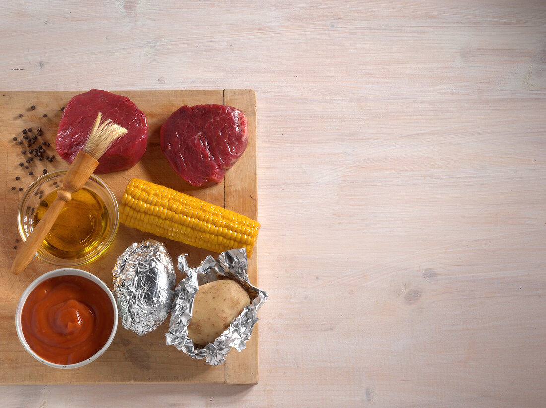 Two steaks, maize cobs, potatoes, barbecue sauce and bowl of oil with brush, copy space