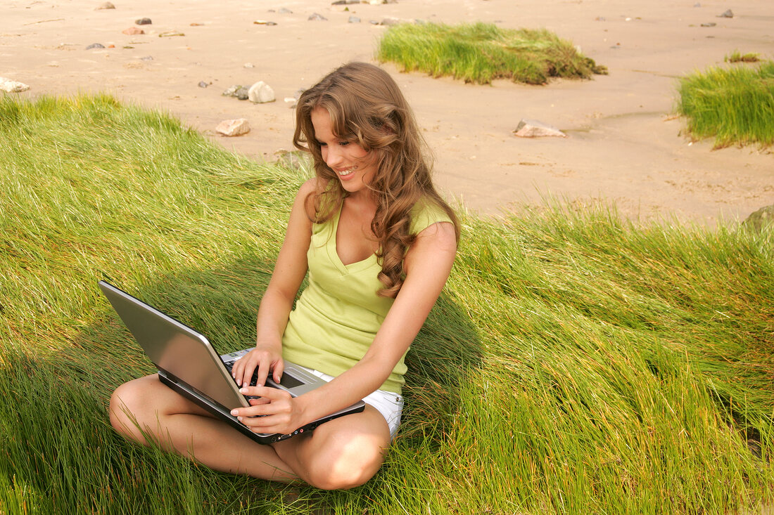 Woman with brown hair wearing green top and white shorts using laptop on beach