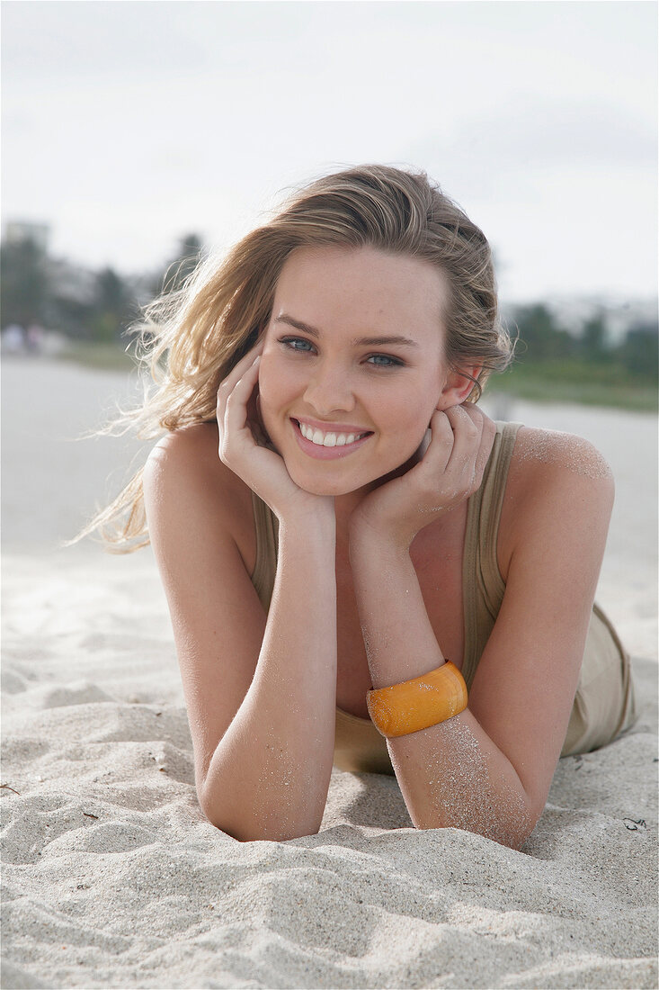 Portrait of beautiful blonde woman wearing brown top lying on beach, smiling