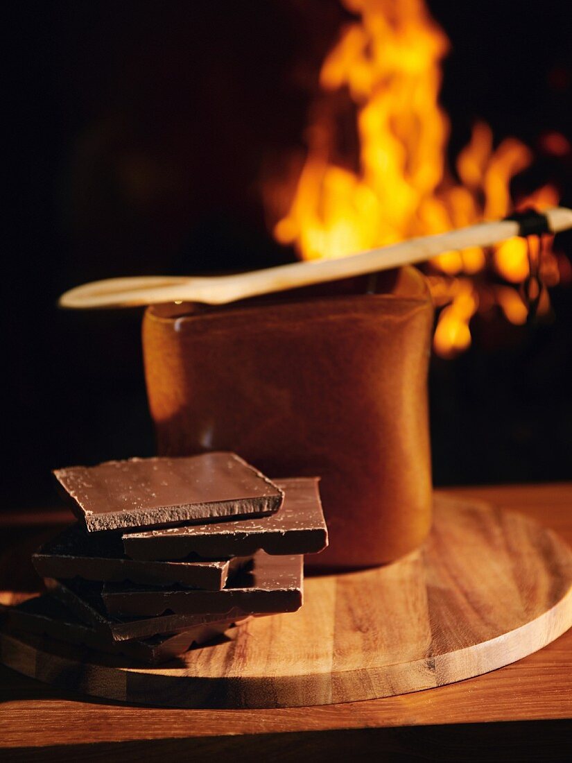 Gestapelte Schokoladenstücke vor Topf, offenes Kaminfeuer im Hintergrund