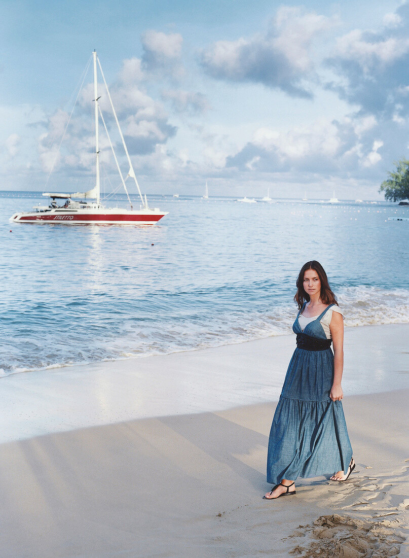 Pensive woman wearing jeans dress walking along beach and boats in background