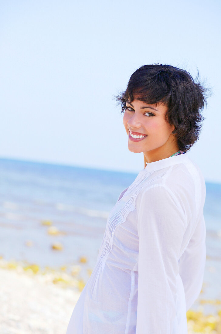 Portrait of brunette woman in white blouse looking over shoulder against sea, smiling