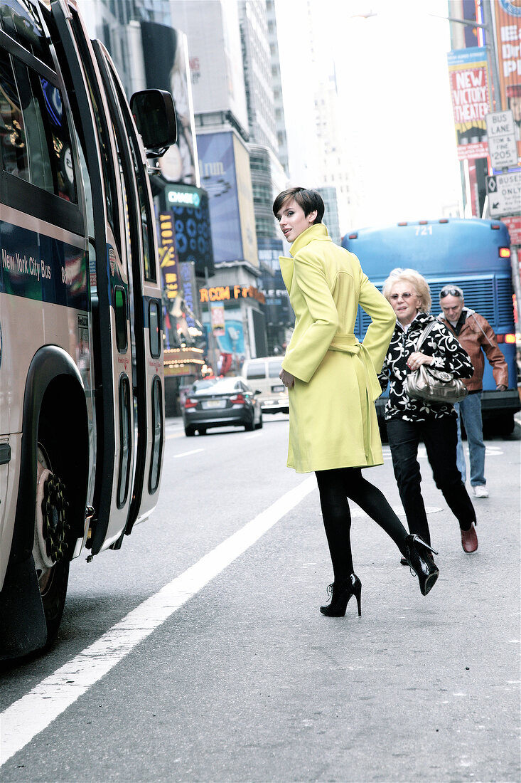 Woman wearing long coat and high heels walking on street along bus, looking over shoulder