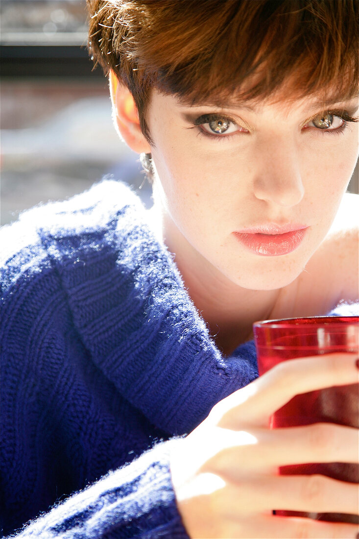 Portrait of green eyed woman with short hair in blue sweater holding red glass with drink
