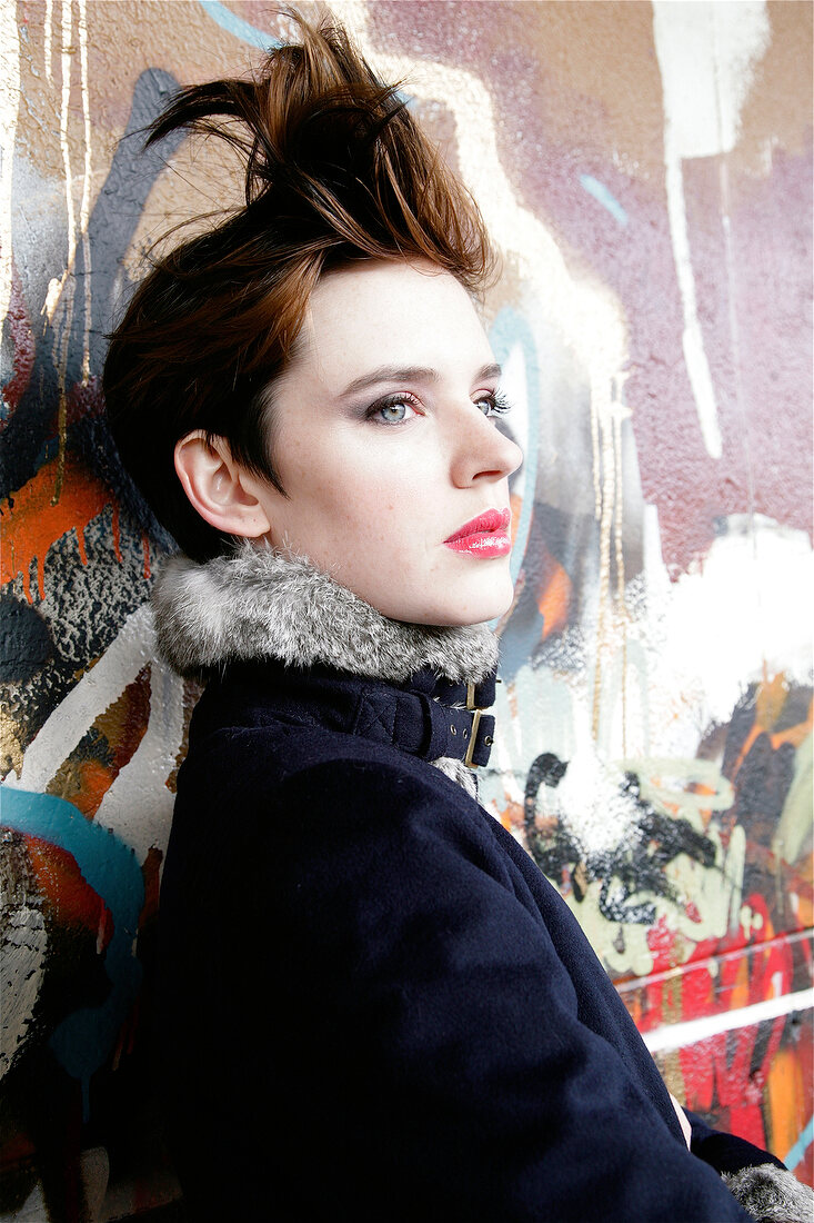 Pretty woman with short hair wearing make-up leaning against wall, looking away