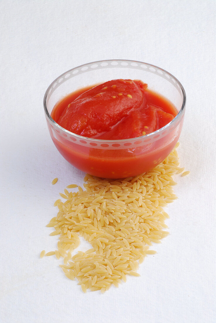 Raw kritharaki and peeled tomatoes in glass bowl