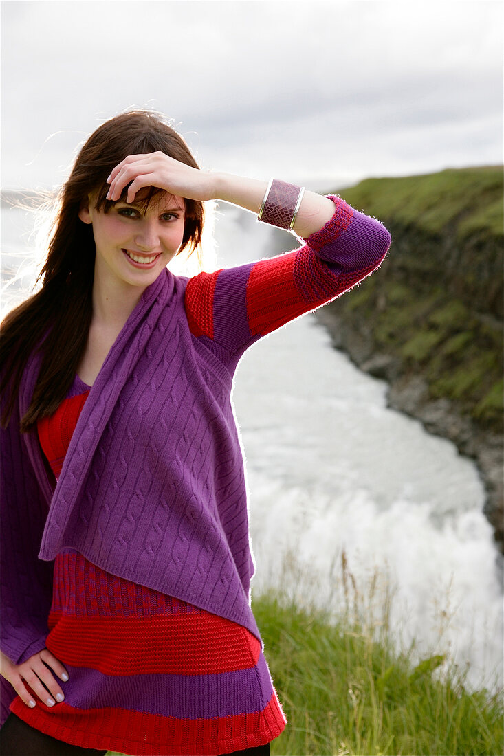 A brunette woman wearing knitwear with her arm to her forehead, stands in front of water