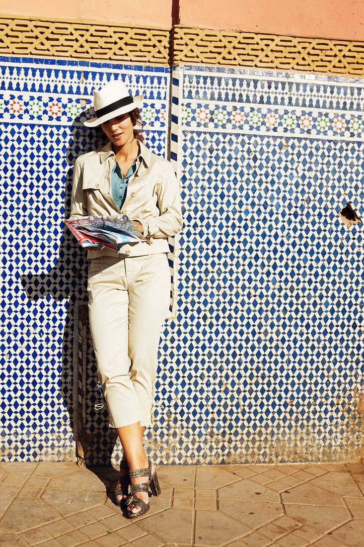 Young woman in beige hat and coat leaning against wall and looking at map of Marrakech