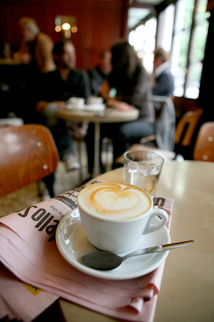 Cup of cappuccino with heart shape on froth, kept on newspaper on cafe table