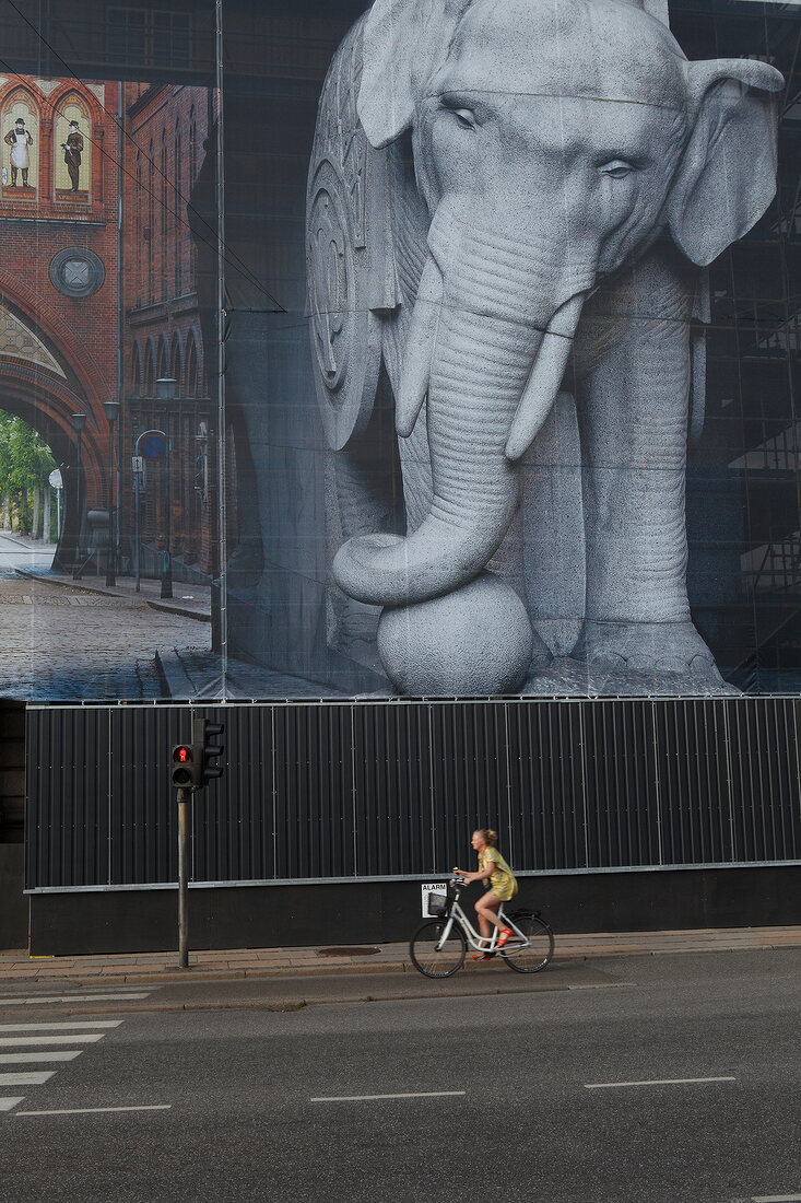 Girl riding bicycle beside building with elephant cladding in Copenhagen, Denmark