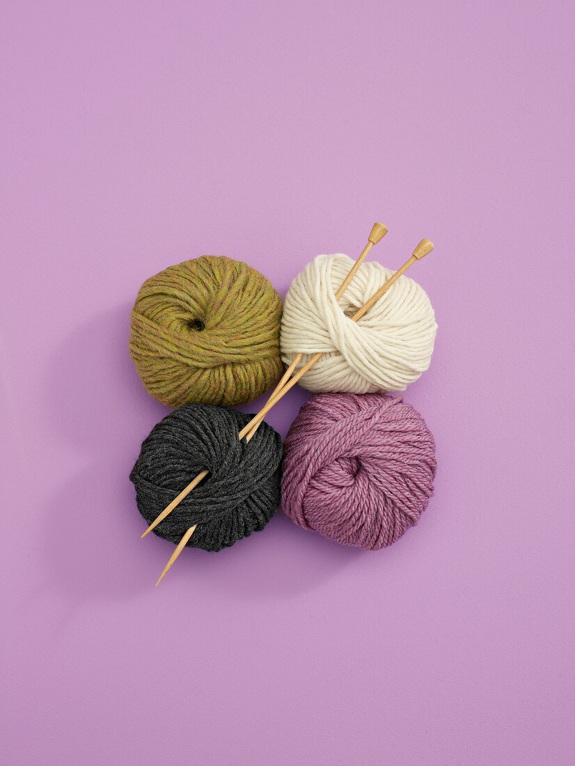 Four balls of wool in different colours with knitting needles on pink background