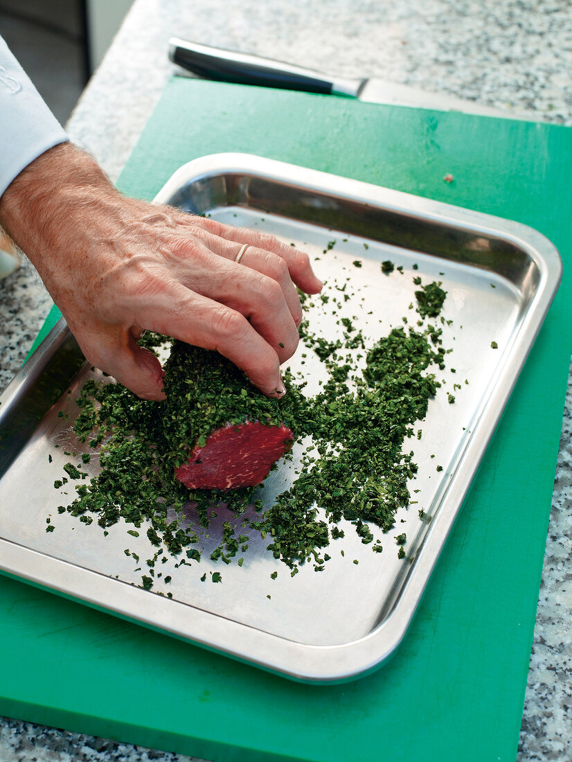 Close-up of man's hand rolling beef in chopped herbs on steel tray