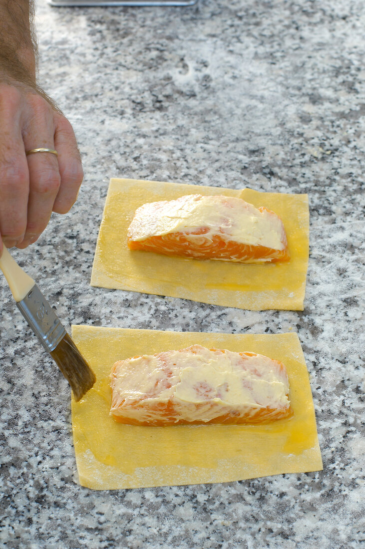 Close-up of man's hand applying butter with brush on salmon fillet on pasta dough
