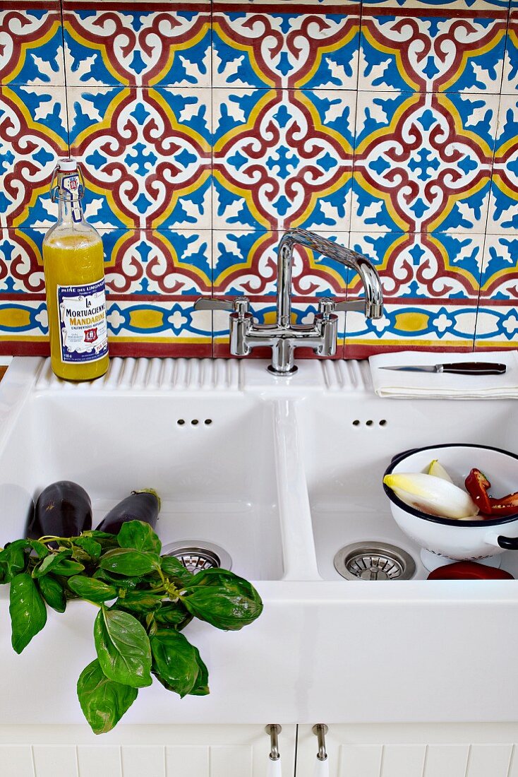 Basil sprigs and vegetables in a white ceramic sink with Moroccan tiles on the wall behind
