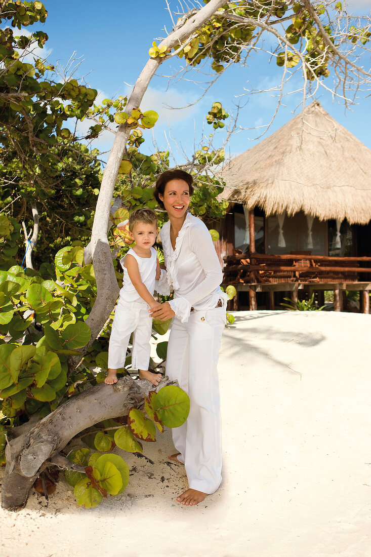 Mother and son in white outfit standing on beach under a tree, smiling