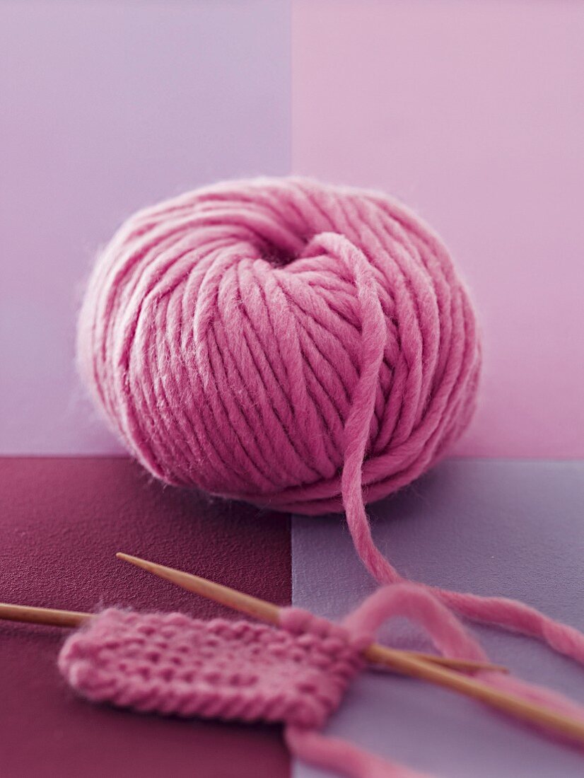 A pink ball of wool with knitting needles on a pink and purple surface