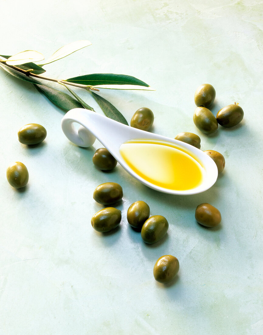 Spoon with olive oil, green olives and olive tree branch on green background