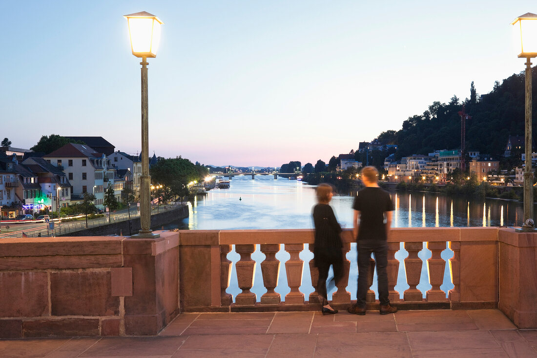 Couple standing on Karl-Theodor Bridge overlooking Neckar at dusk in Heidelberg, Germany