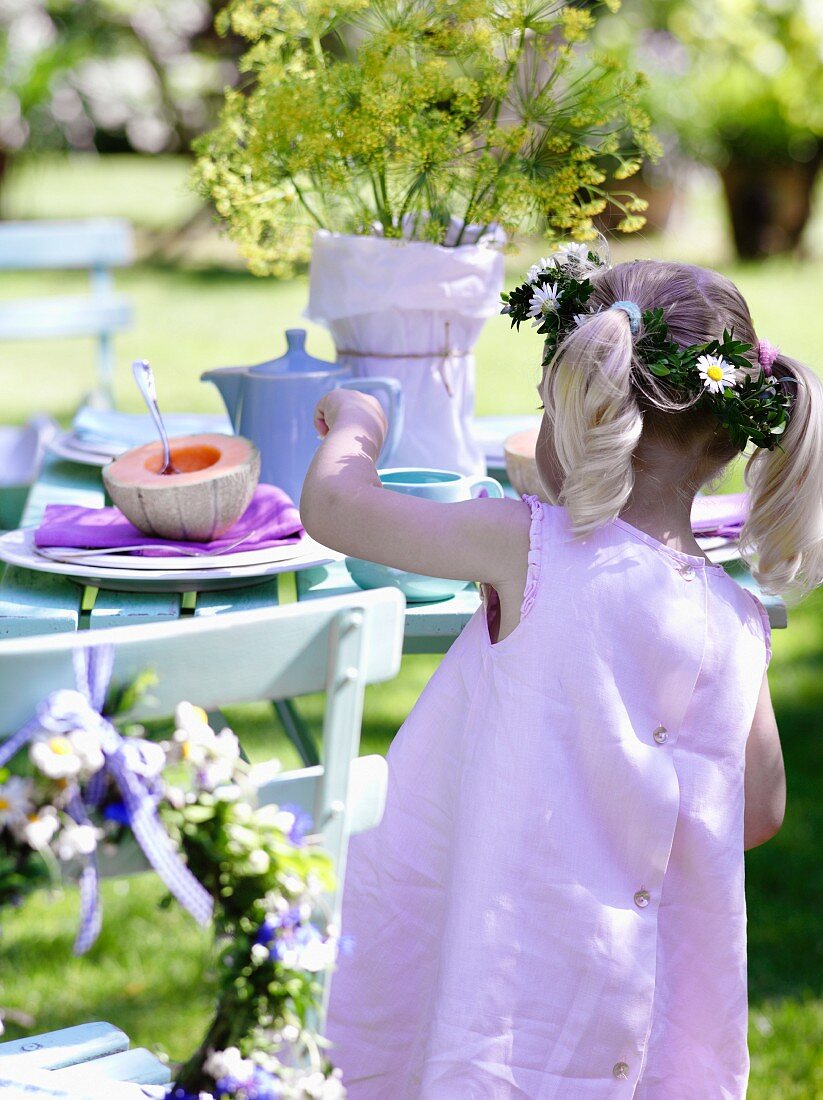 A girl wearing a pink dress and a crown of flowers by a table in a garden