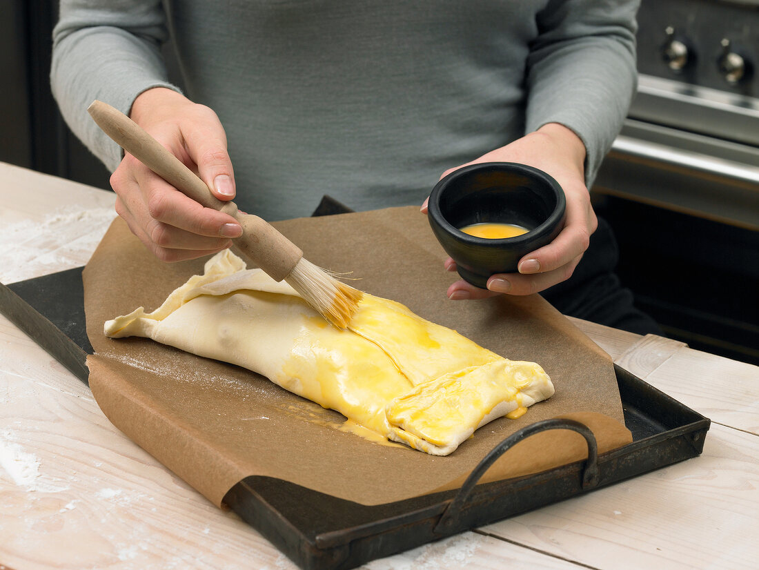 Hand applying egg yolk with brush on apple strudel in baking dish, step 3