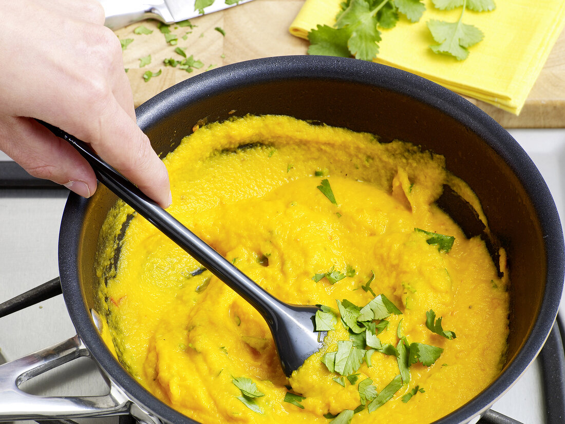 Close-up of hand mixing carrot coulis and herbs with spoon in sauce pan, step 5