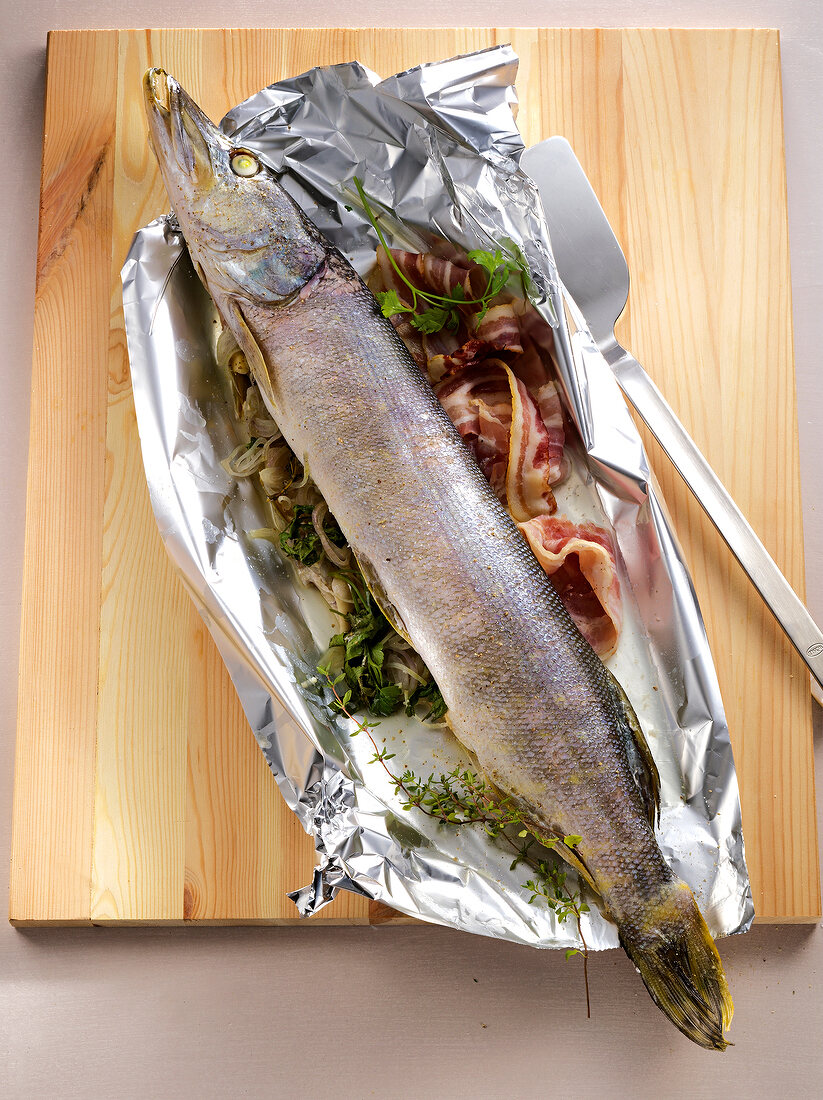 Northern pike wrapped in bacon in silver foil on wooden board