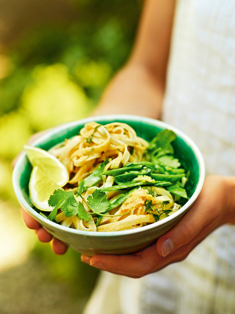 Woman holding bowl of rice noodles with snow peas and lemon, garden kitchen