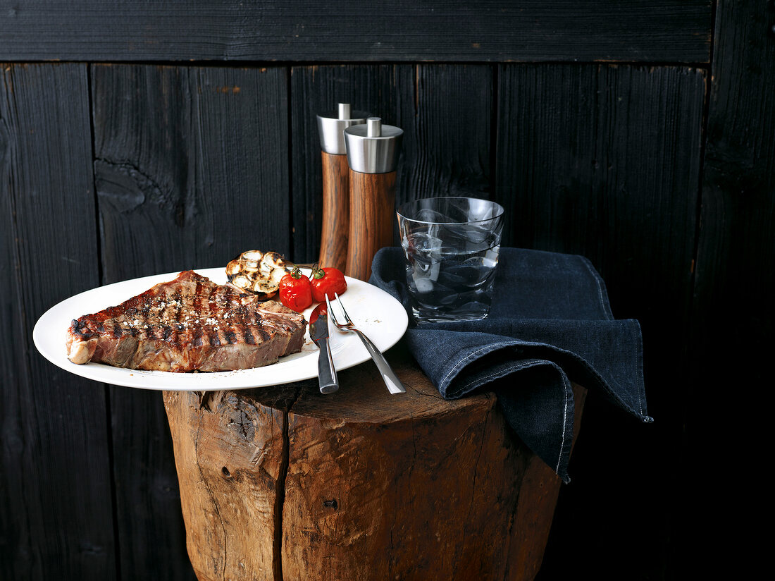 Plate of grilled steak with glass of water, napkin, salt and pepper mill on wooden block