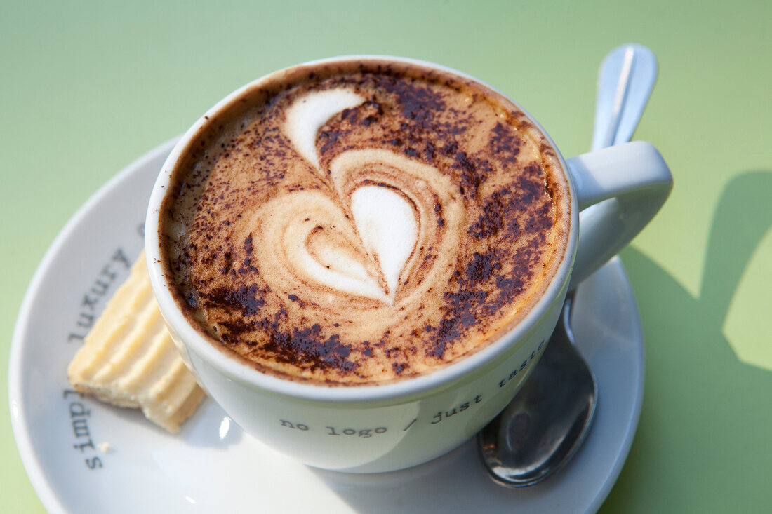 Close-up of coffee with heart shaped cream design in cup with spoon and cookie on plate