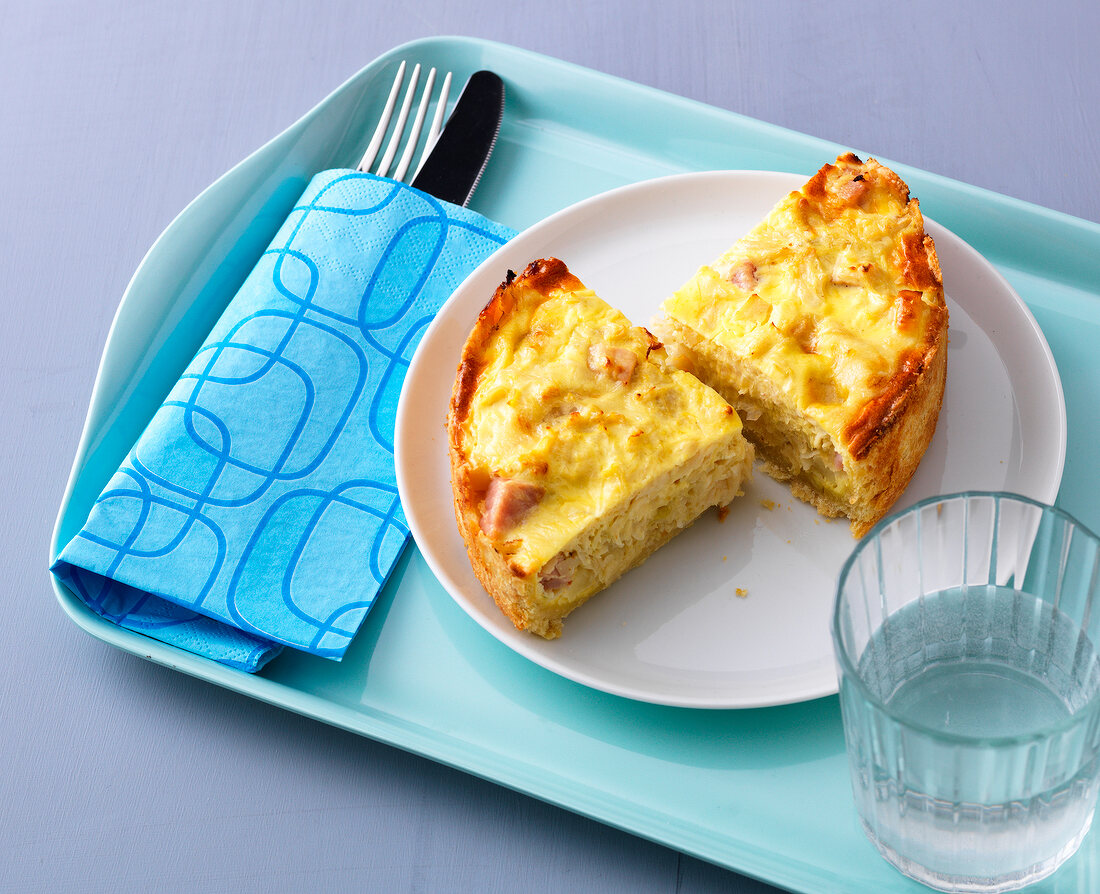 Two pieces of quiche on plate with glass of water and cutlery on tray