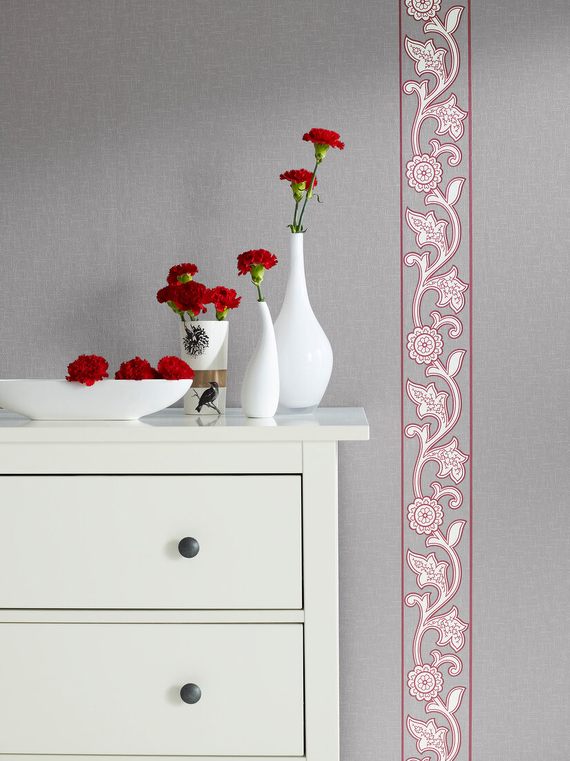 White flower vase on chest of drawers in front of gray tapet with Paisley patterned border