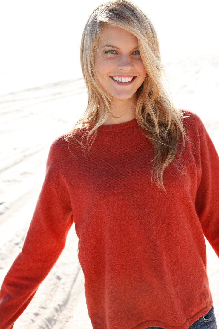 blonde Frau in orangefarbenem Pulli und Jeans am Strand