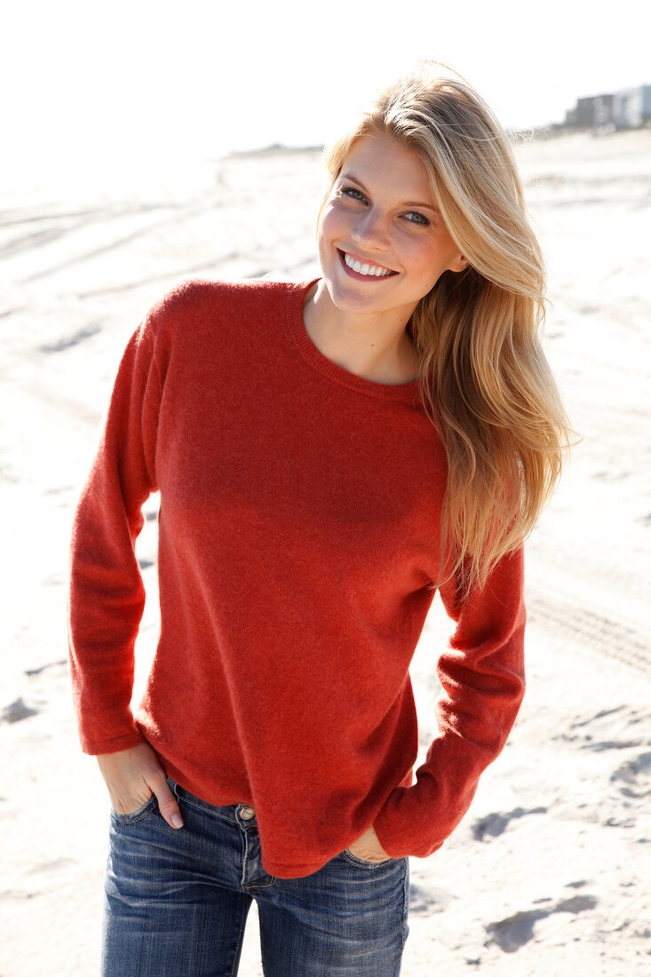 Pretty blonde woman in orange top and jeans standing on beach, smiling