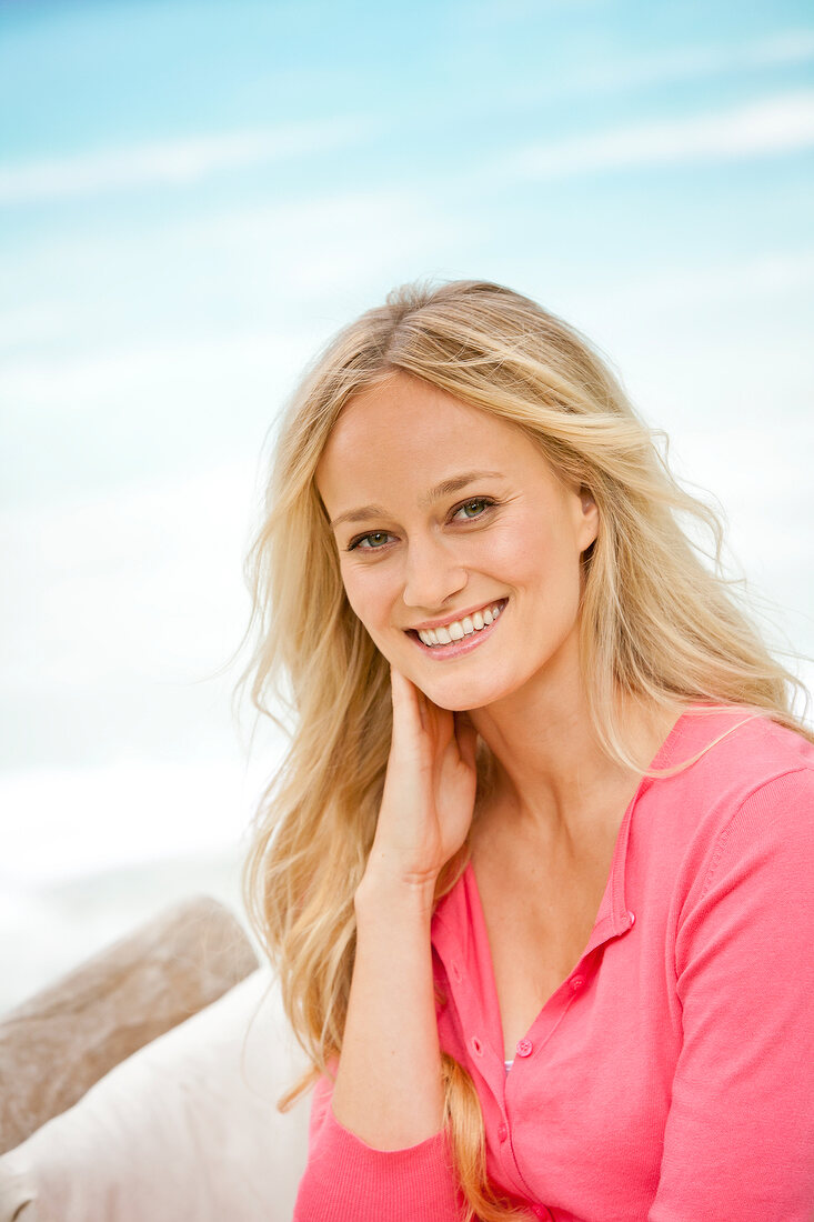 Portrait of beautiful blonde woman wearing pink sweater sitting and smiling on beach