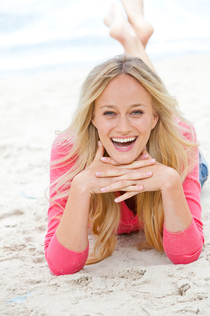 Portrait of happy blonde woman wearing pink sweater lying on sand and smiling
