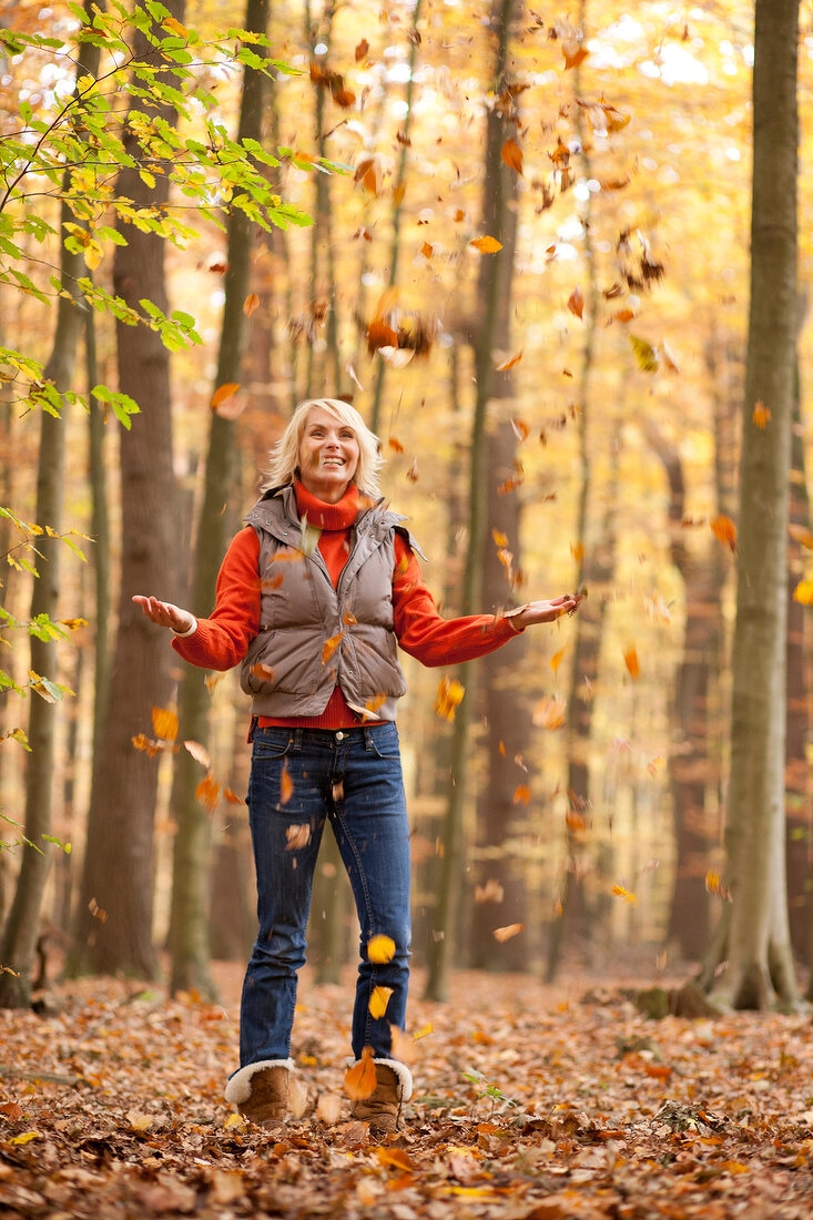 Happy blonde woman wearing gray jacket and jeans playing with falling leaves, smiling