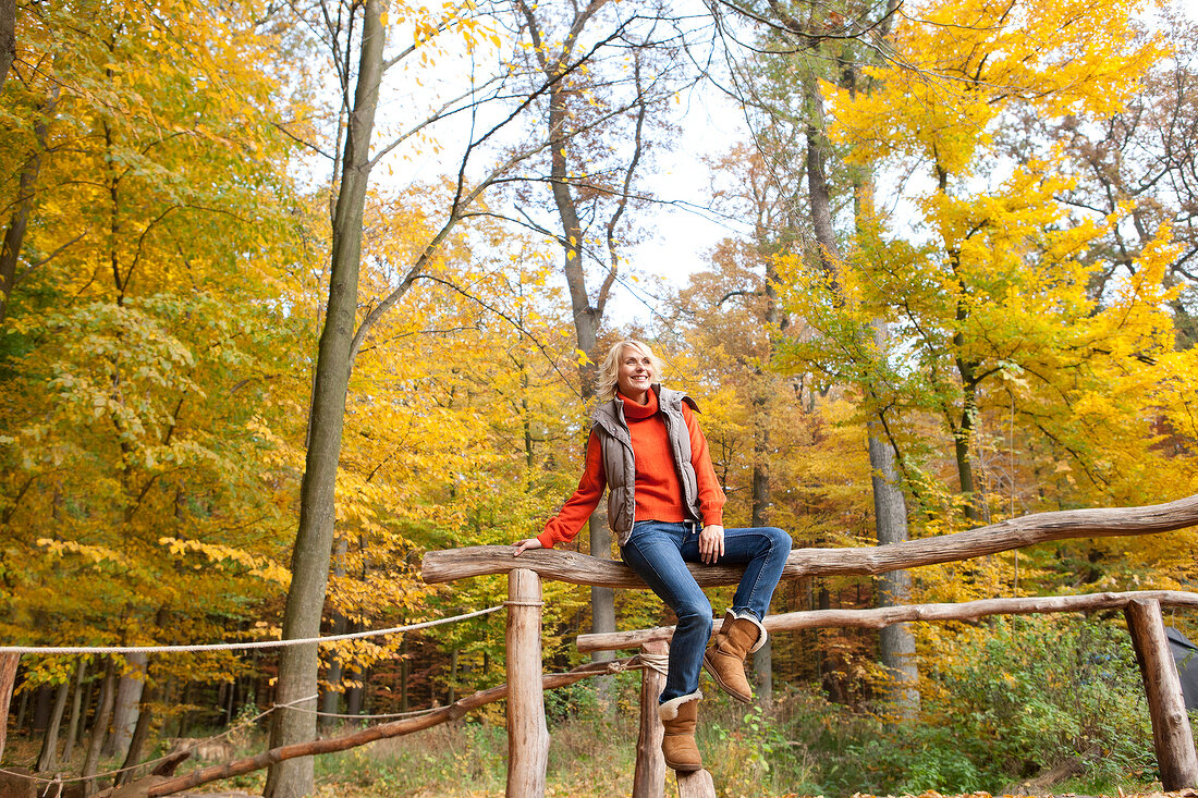 Blonde woman sitting on a wooden fence in autumn forest, smiling