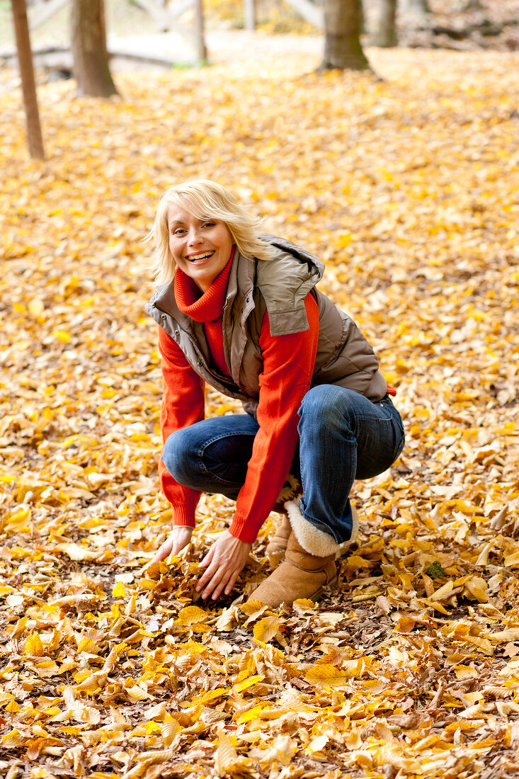 Happy blonde woman in gray jacket sitting and collecting autumn leaves in forest, smiling