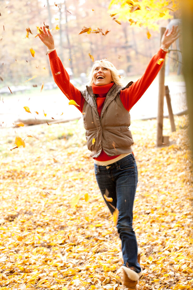 Excited blonde woman wearing gray jacket and jeans playing with autumn leaves in forest