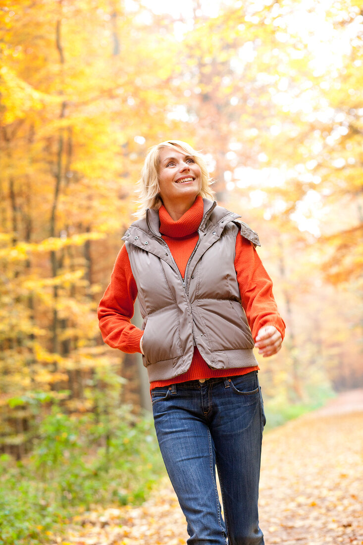 Blonde woman wearing gray jacket and jeans looking upwards while walking in autumn forest