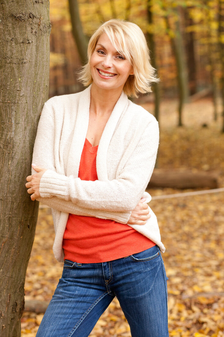 Blonde Frau in beigefarbener Jacke lehnt an einem Baum