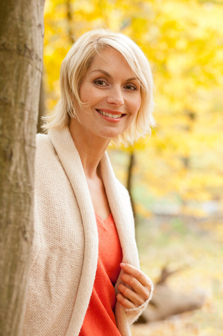 Pretty blonde woman with short hair in white sweater standing beside tree, smiling