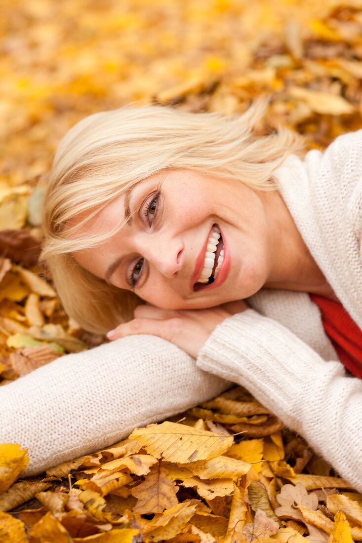 Close-up of pretty blonde woman wearing white sweater lying on autumn leaves