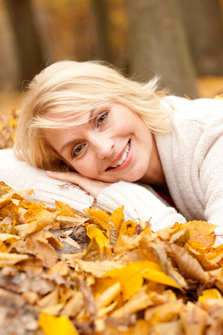 Portrait of happy blonde woman wearing white sweater lying on autumn leaves, smiling