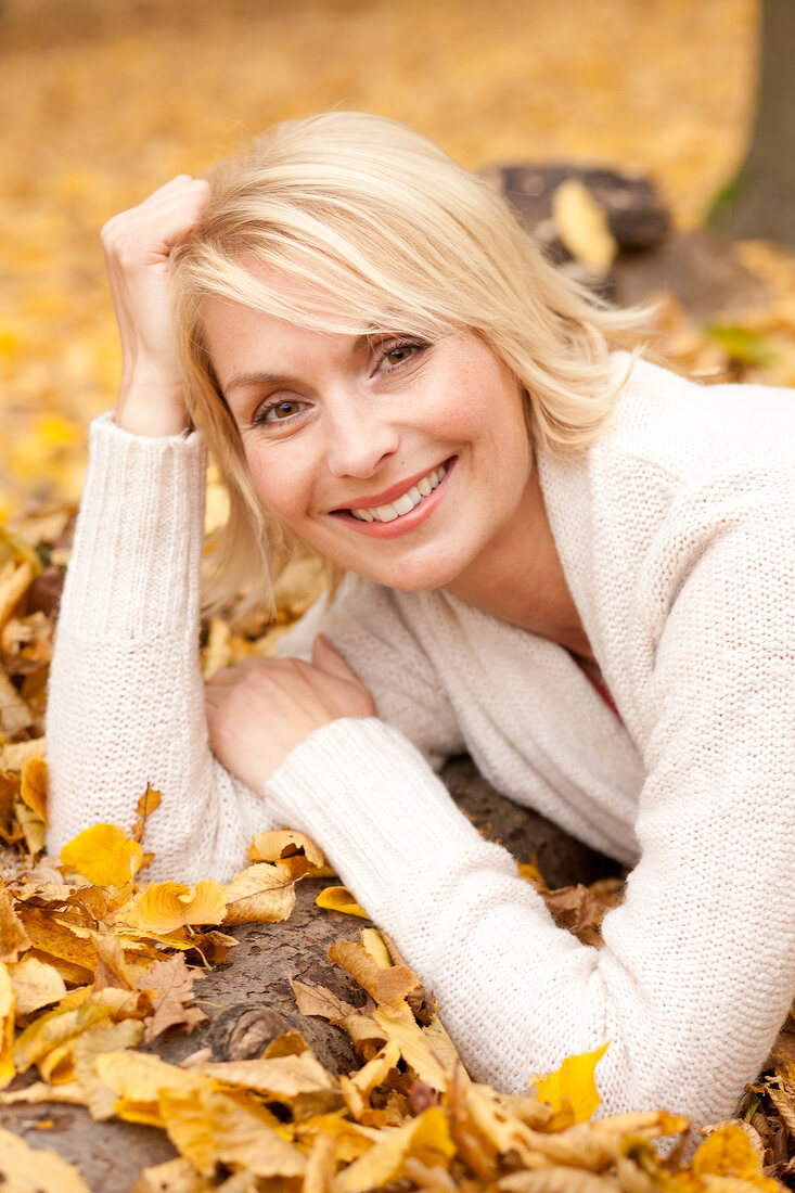 Pretty blonde woman wearing white sweater lying on autumn leaves with head on her hand
