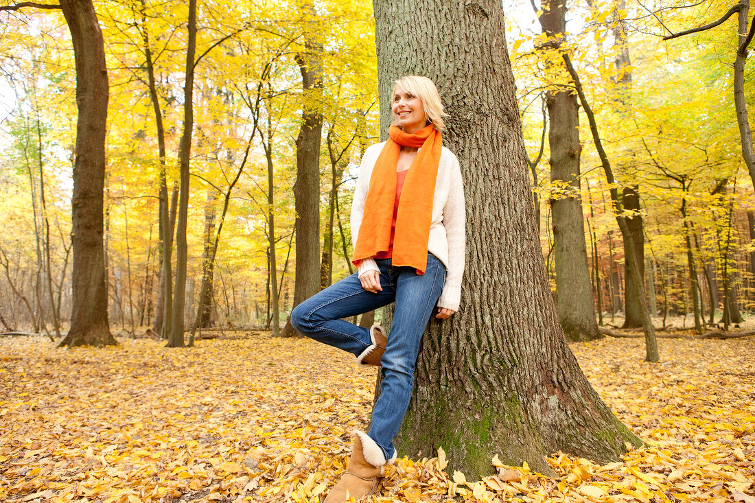 Blonde woman wearing scarf and cardigan leaning against tree in autumn forest, smiling