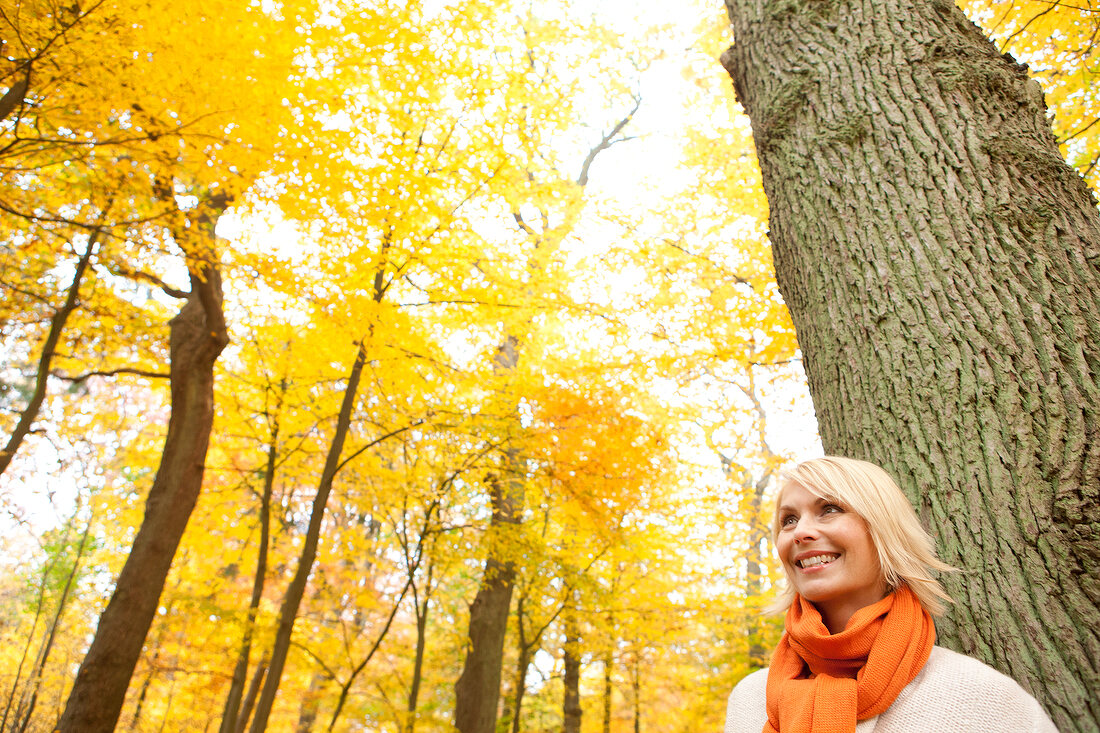 Blonde woman wearing orange scarf leaning against tree in autumn forest, smiling