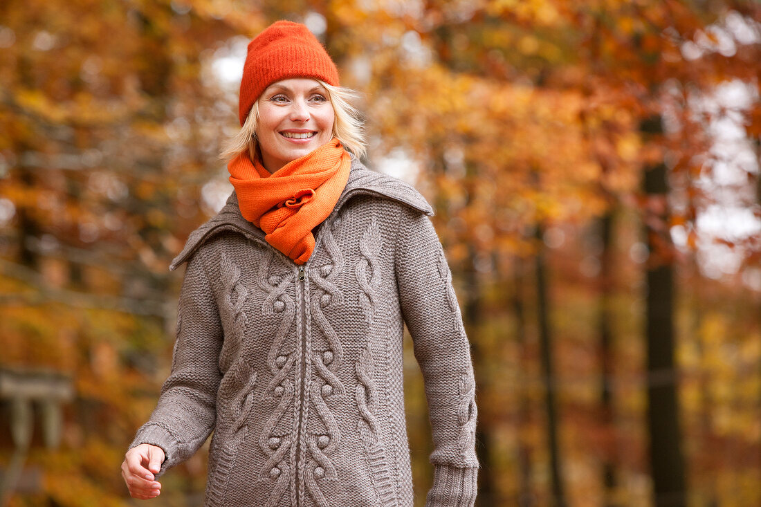 Cheerful blonde woman in gray sweater, orange scarf and cap, smiling in autumn forest