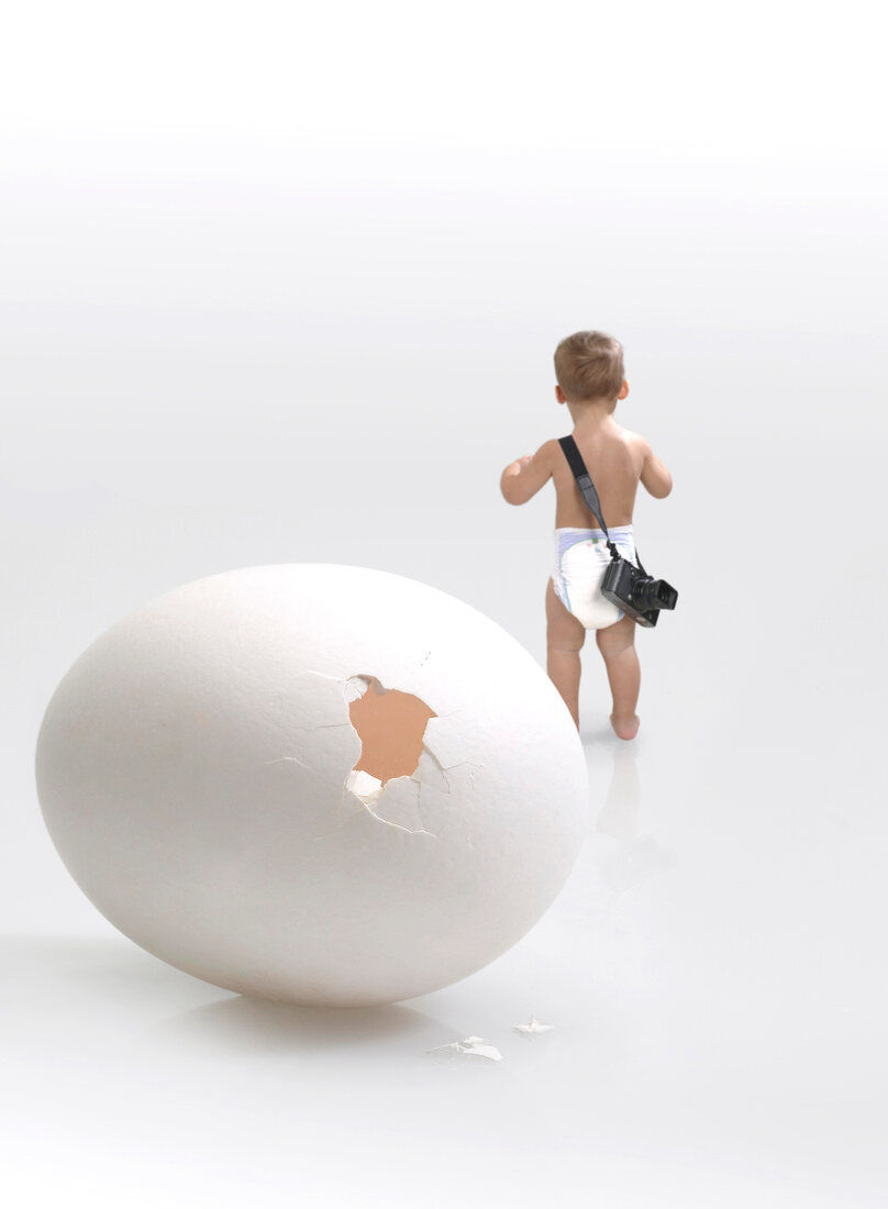 ein kaputtes weißes Ei, Baby in Windeln von hinten mit Kamera
