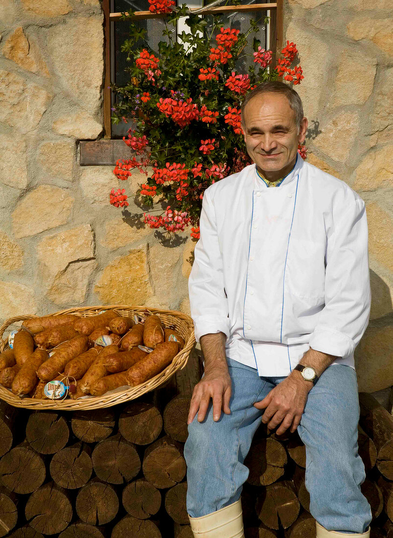 Man sitting on pile of woods with basket full of sausages beside him, France